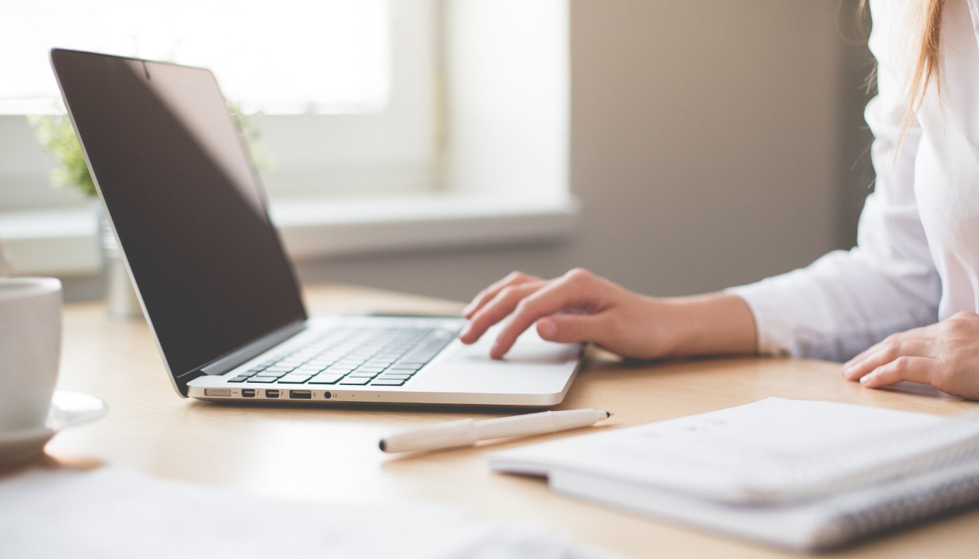 IT Support Technician header image of a woman using a laptop with a notebook and pen on the desk beside her